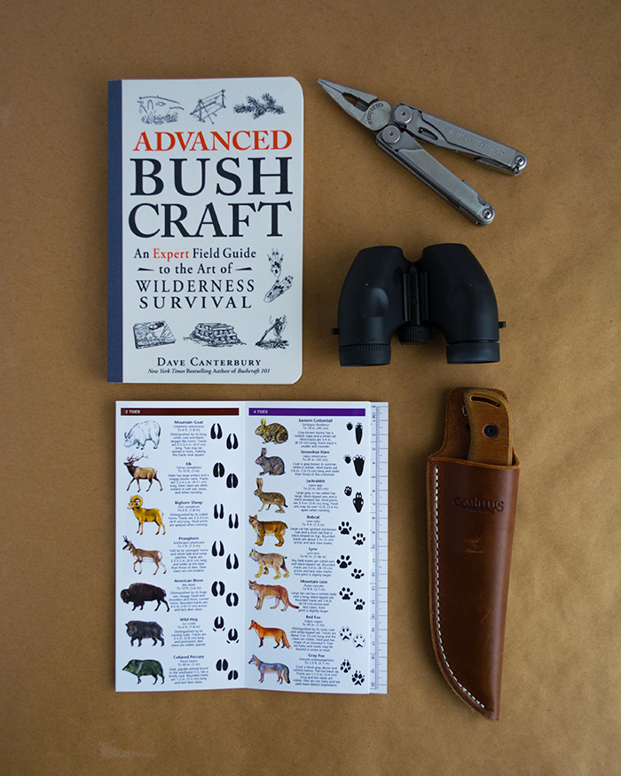 Photo flat-lay with an advanced bushcraft book, leatherman, binoculars, knife in a leather sheath, and a card to help identify animal tracks.
