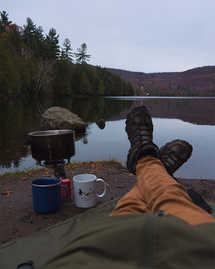 Woman's legs and feet extended with a propane camp stove on the left side and a lake and hills in the background