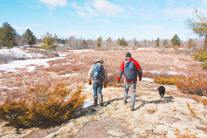 Two men are hiking with their backs to the camera across frozen ground. The sky is blue and the landscape is red-brown with snow.