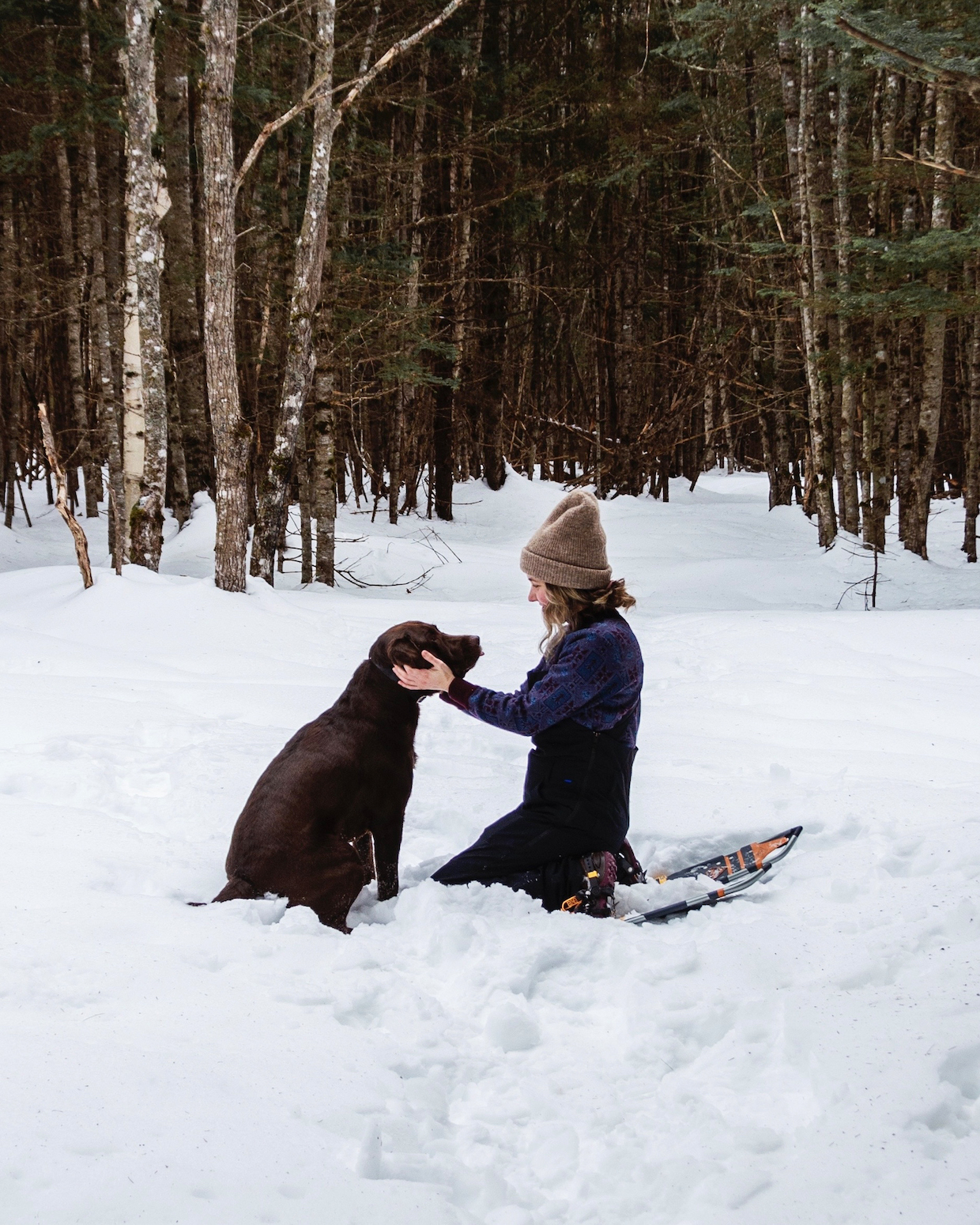 Jen and her dog, Moose, on a snowshoe together. She bends down on her knees to pet him. Solo hiking, or snowshoeing, can be less lonely with a dog.