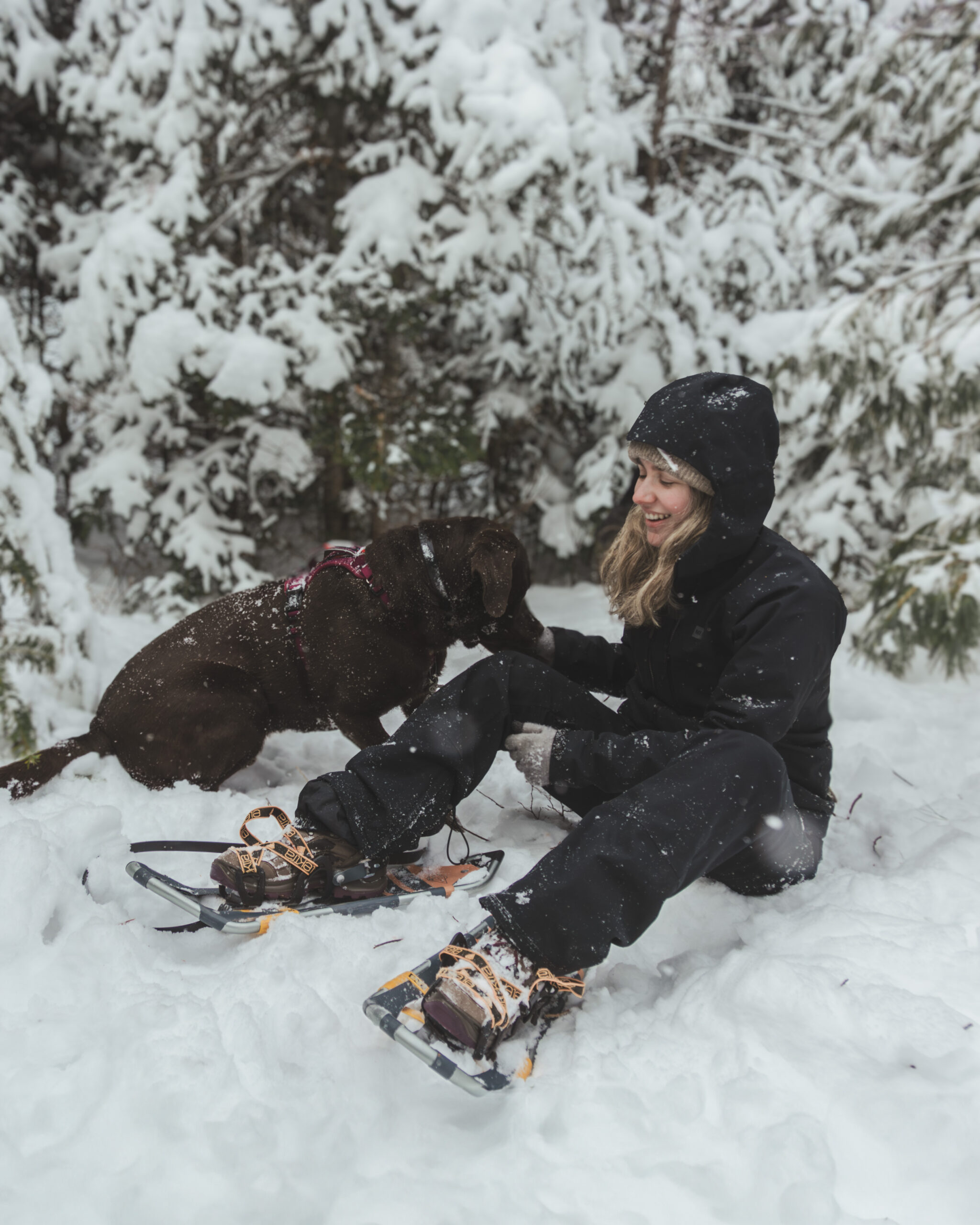 A woman sits in the snow with her chocolate lab. She's wearing snowshoes and her hood is pulled over her head. A row of snow covered conifers are in the background.